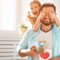 A man and young girl share a gift box indoors, with a card featuring a heart symbol, representing family joy.