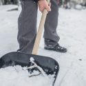 Person shoveling snow on a snow-covered ground with a wooden-handled shovel, illustrating winter home maintenance tasks.