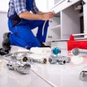 Person in blue overalls working on plumbing under a cabinet with tools and fittings on the floor for home maintenance.