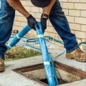 Person in work attire using plumbing equipment, including a blue hose and drain cover, showcasing hands-on maintenance work.
