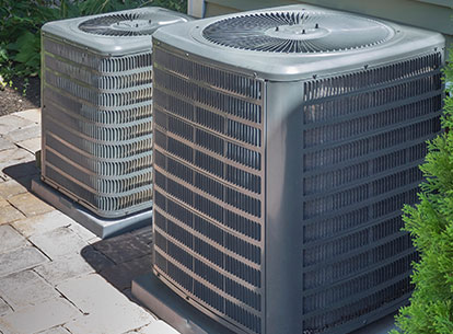 Two outdoor air conditioning units positioned on a stone patio, surrounded by greenery, representing cooling services offered by Laney's Inc. for home comfort.